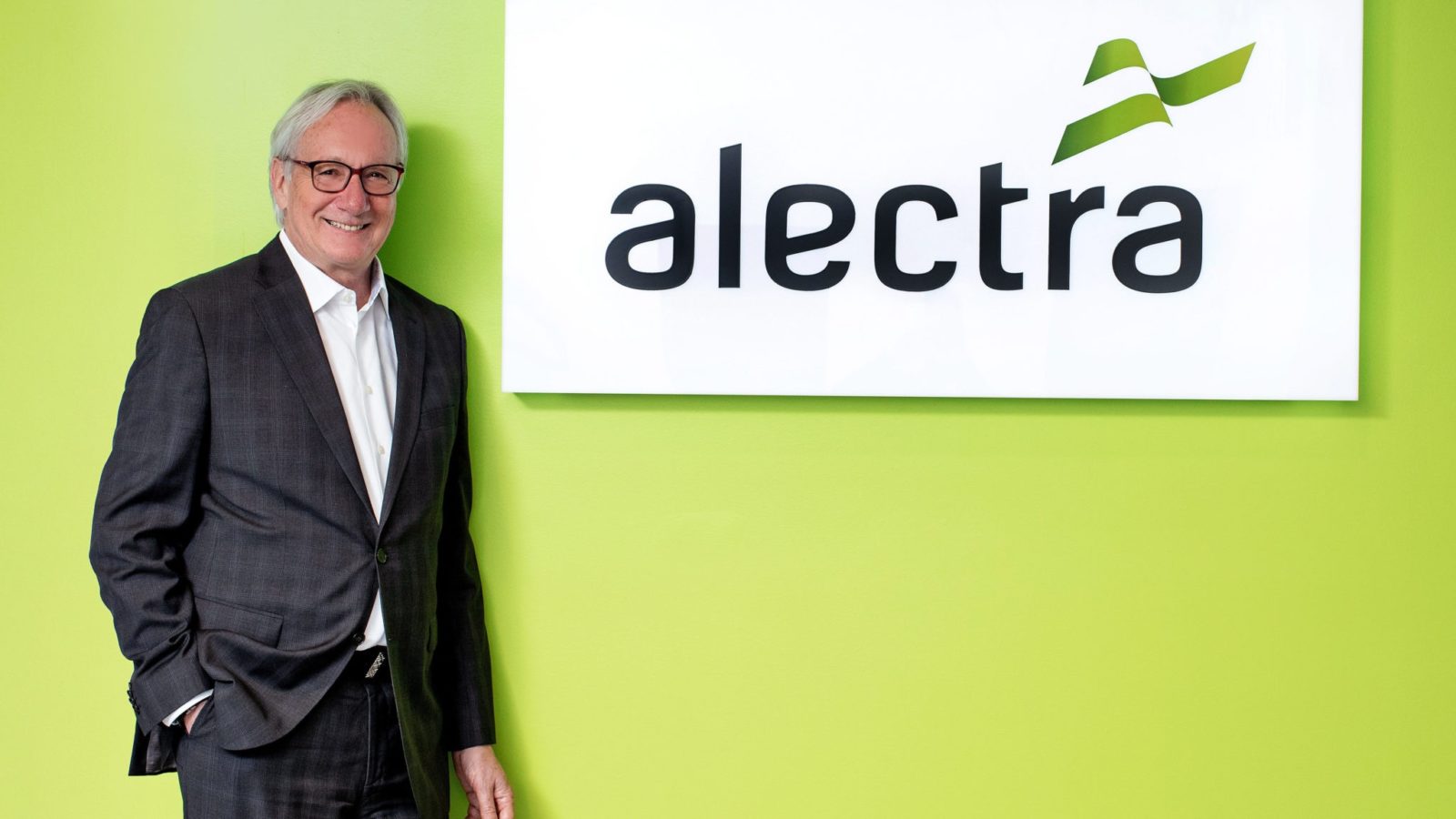 Blair Peperdy, with his hand in his pocket and wearing a black suit, stands beside an alectra logo and smiles