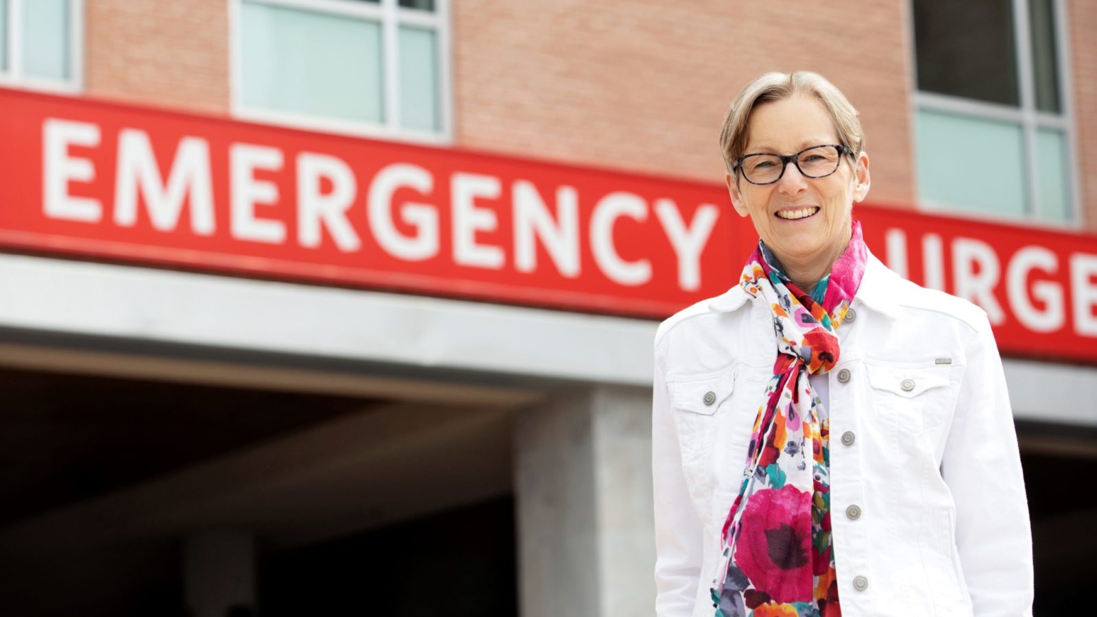 Karen Henkenhaf, wearing a floral scarf and white denim jacket, stands outside the RVH emergency sign and smiles for a photo