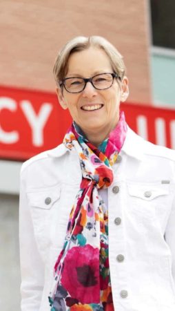 Karen Henkenhaf, wearing a floral scarf and white denim jacket, stands outside the RVH emergency sign and smiles for a photo