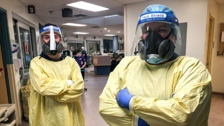 two nurses in yellow protective equipment and face shields stand together with arms crossed