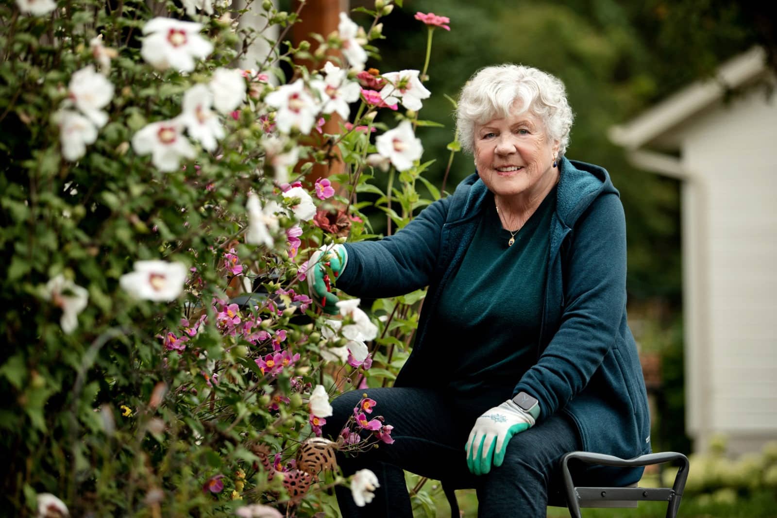 Judy Carson sits next to her garden, smiling, wearing gloves and using sheers to prune her blooms.