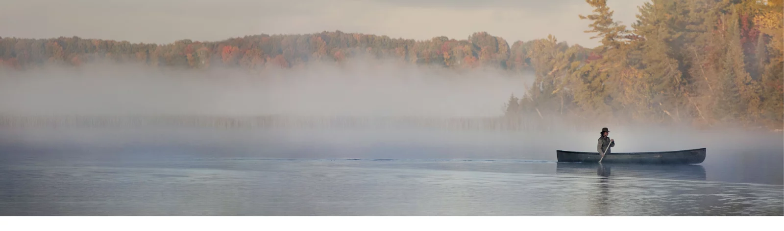 Drew Hodgins paddles a canoe on a calm lake. Fog over the water blurs the line between lake and sky.