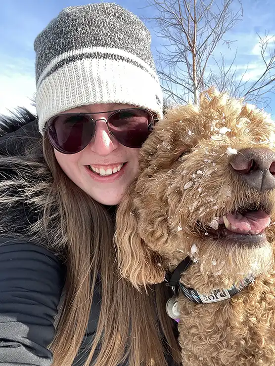 Michelle Menzies, wearing a white hat, sun glassses, and black coat, while taking a selfie with her doodle