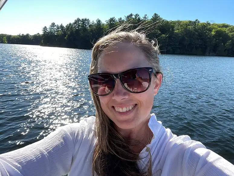 Robyn Blanchet, seen from the shoulders up, wearing a white shirt and sunglasses. A lake and some trees can be seen in the background